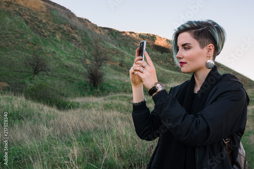 Young woman take a picture on smart phone in the green field with hillon background during spring or autumn time photo