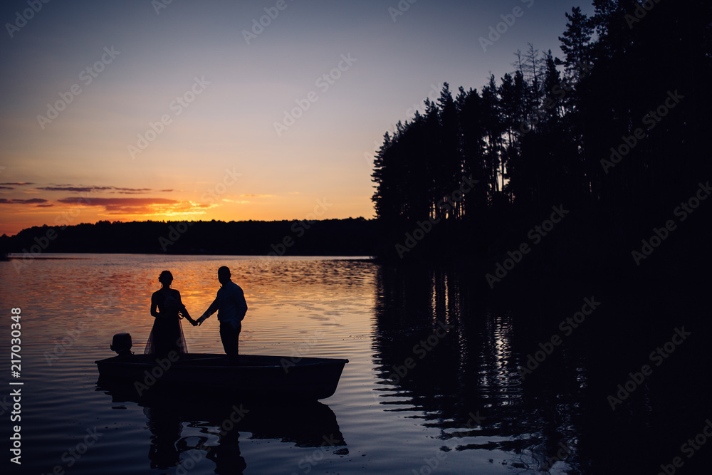 Silhouette of two lovers in boat on the lake at sunset. Photosession of couple on water Silhouette of two lovers in boat on the lake at sunset. Photosession of couple on water