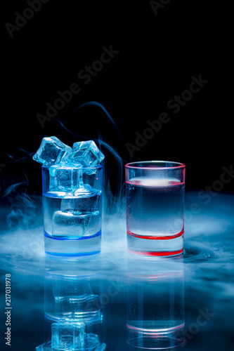 Two, blue and red shot glasses full of drinks one of wich with ice in a theatrical smoke on a black background.