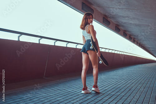 Back view of a sensual young skater girl dressed in shorts and t-shirt holds a skateboard and looking at the camera.