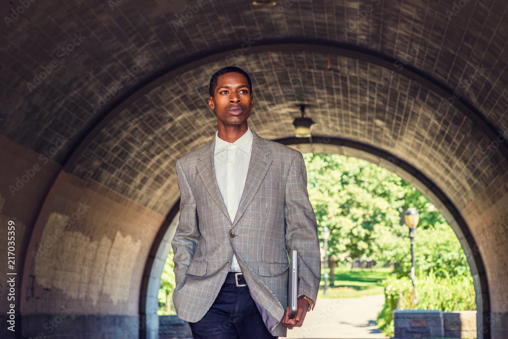 Young African American Businessman traveling in New York, wearing patterned blazer, white undershirt, black pants, holding laptop computer, walking through under street bridge, looking forward