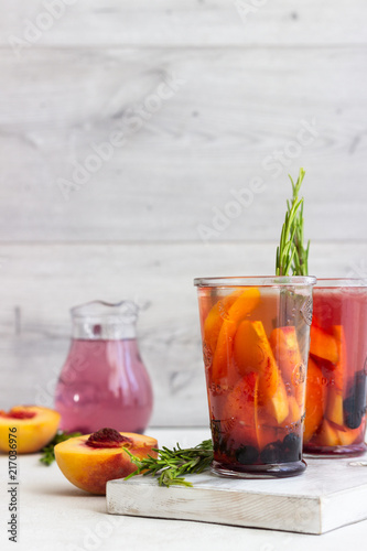 Peach and rosemary lemonade or cocktail in glass with fresh peaches on a light gray background. Summer cold drink.