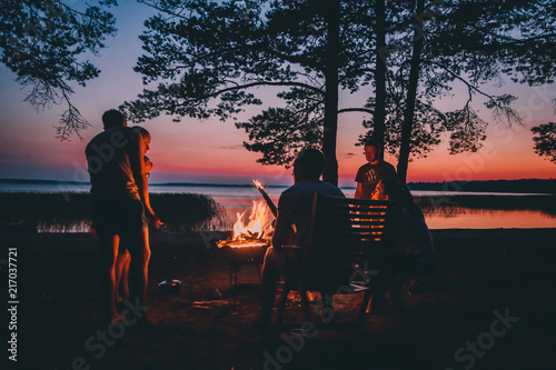 Holiday with friends in nature. Group of young people near camp fire , telling stories near the fire with wood, flames in the nature at night near lake.