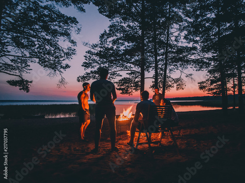 Holiday with friends in nature. Group of young people near camp fire , telling stories near the fire with wood, flames in the nature at night near lake.