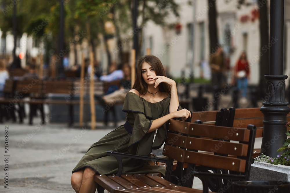Sensual and fashionable brunette model girl in stylish dress with naked shoulders sits on a bench and posing outdoors at the walking street Sensual and fashionable brunette model girl in stylish dress with naked shoulders sits on a bench and posing outdoors at the walking street