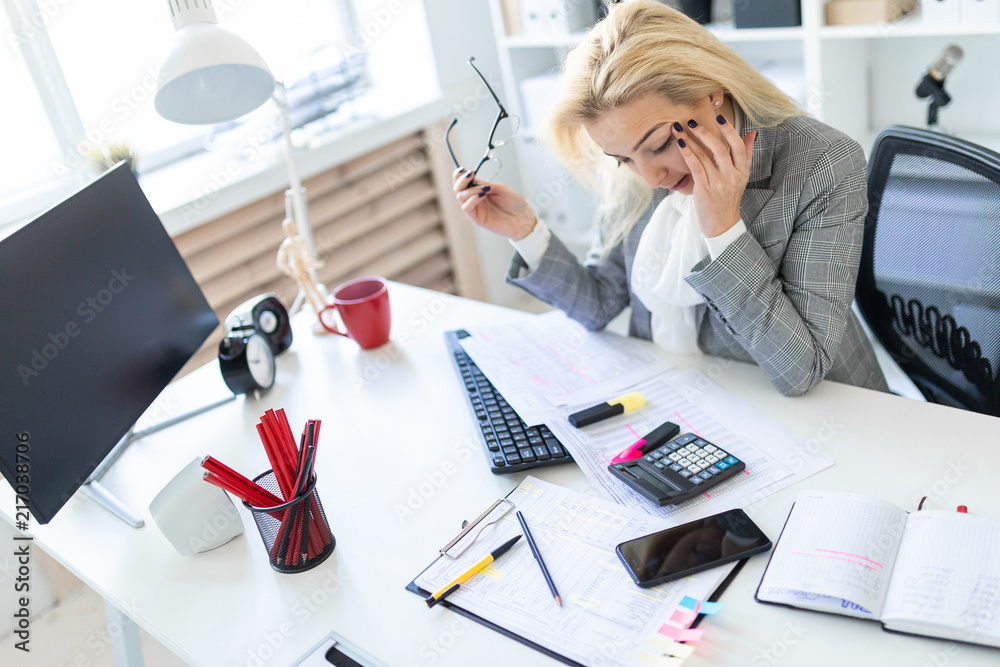 A young girl is sitting at a table in the office. The girl is holding glasses and rubbing her eyes. A young girl is sitting at a table in the office. The girl is holding glasses and rubbing her eyes.