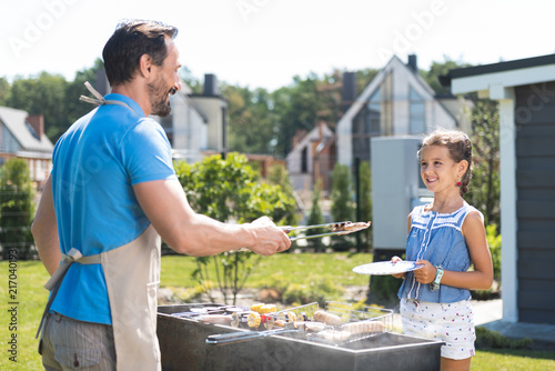 Grilled food. Positive nice man looking at his daughter while putting sausages on her plate