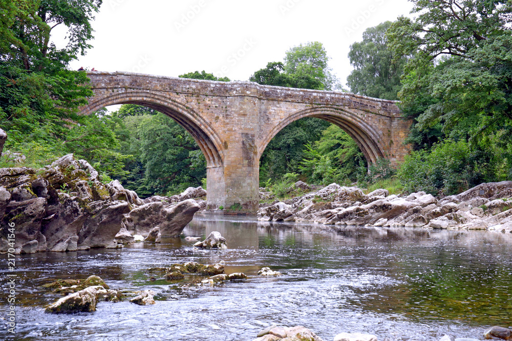 The Devil’s Bridge Kirkby Lonsdale Cumbria, an historic gritstone bridge over the River Lune.Landscape with a foreground of limestone rocks and river flowing under the bridge