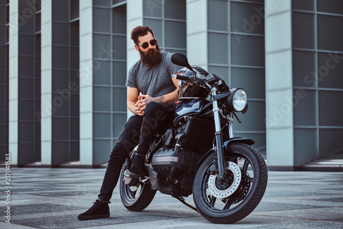 Brutal bearded male in sunglasses dressed in a gray t-shirt and black pants sitting on his custom-made retro motorcycle against a skyscraper.