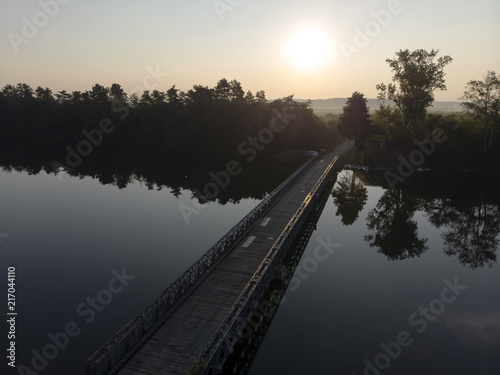 Old wooden bridge with rails over river against sunset background.