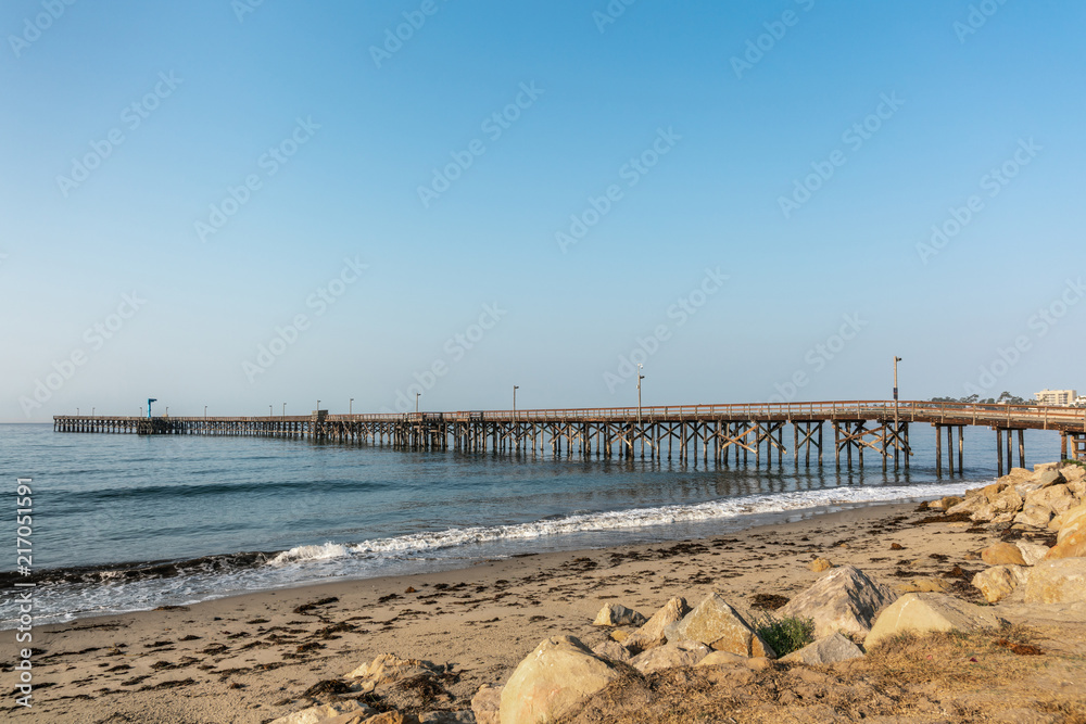 Goleta, California, USA - August 7, 2018: Long wooden pier stretches far into calm Pacific Ocean from right to left under blue sky. Sandy beach up front. 