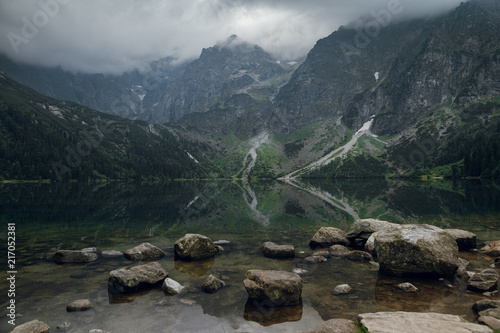 Scenic view of beautiful foggy rocky mountains and green forest with a reflection in a lake. Stony shore on Morskie Oko. Travel concept. High Tatras, Poland