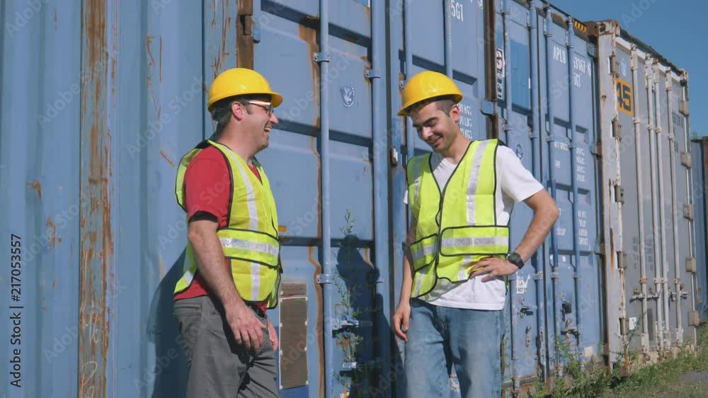Two workers chatting during break time at work in a shipping yard filled with large shipping containers. Two workers chatting during break time at work in a shipping yard filled with large shipping containers.