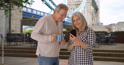 Mature Caucasian husband and wife look at photos on phone near Tower Bridge