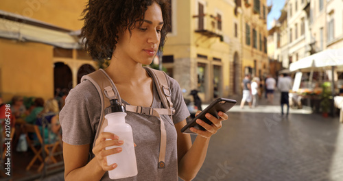 Photography Cute black female tourist texts on smartphone holding water bottle on street in