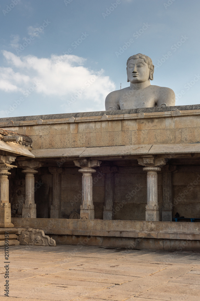 Shravanabelagola, Karnataka, India - November 1, 2013: At the Jain Tirth, gray granite Giant Bhagwan Bahubali puts torso above brown stone temple building against light blue sky.