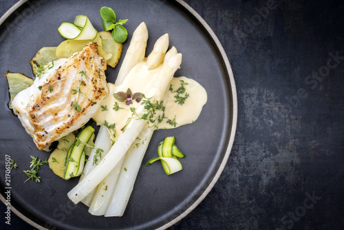 Modern style German fried cod fish filet with white asparagus in hollandaise sauce with roast potatoes and sliced zucchini as top view on a plate with copy space right photo