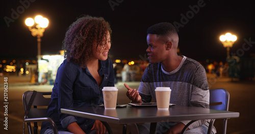 Black man and woman meeting for coffee at night chat and use technology