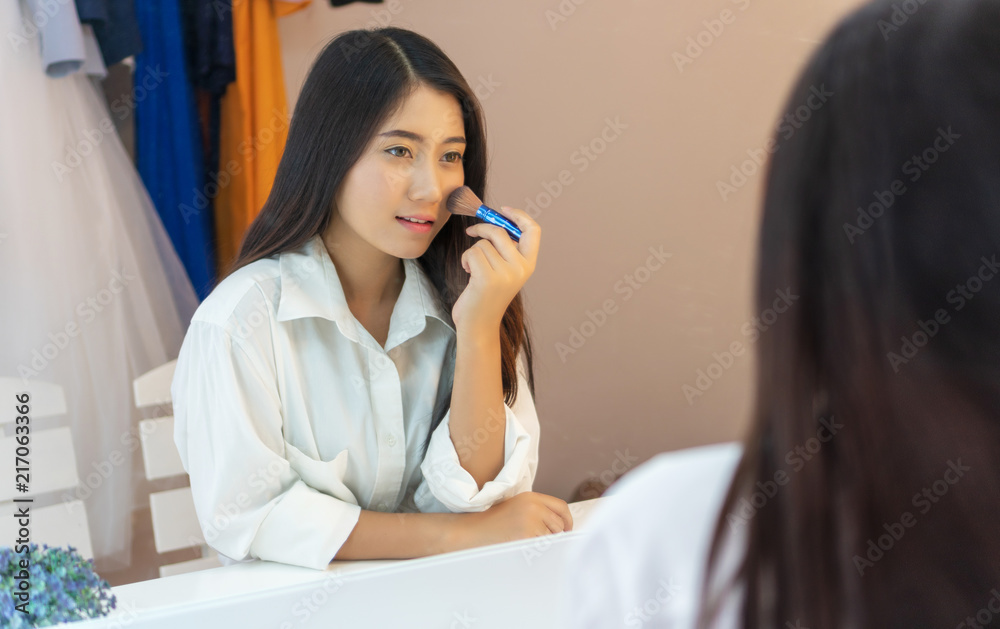 Happy young woman sitting front of mirror doing make – up in the room. Attractive asian woman holding brush and making make up while sitting in front of mirror. Happy young woman sitting front of mirror doing make – up in the room. Attractive asian woman holding brush and making make up while sitting in front of mirror.