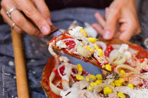 Woman hands hold a piece of raw vegan pizza with dehydrated dough, raw tomato sauce, vegan mayonnaise, corn and fresh vegetables. Healthy Italian food