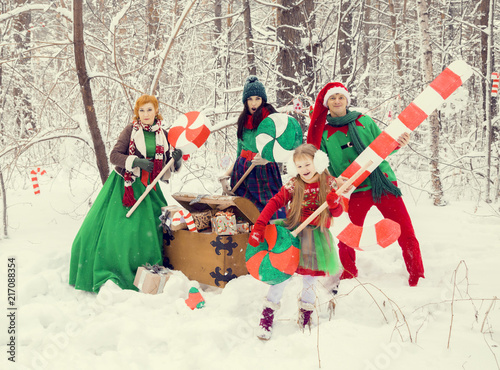 family in traditional Christmas flower costumes, red - green , Santa's helpers - elves, in the winter forest with a chest of gifts and a huge candy