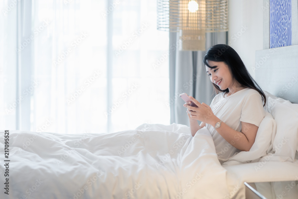 Beautiful young smiling Asian woman lying in white bed and using a phone in her bedroom.. Beautiful young smiling Asian woman lying in white bed and using a phone in her bedroom..