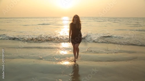 A young sexy blonde woman with long hair in a black short translucent dress and sunglasses playfully enters and leaves the sea circling in the oncoming waves and raises her hands to the sunset. 