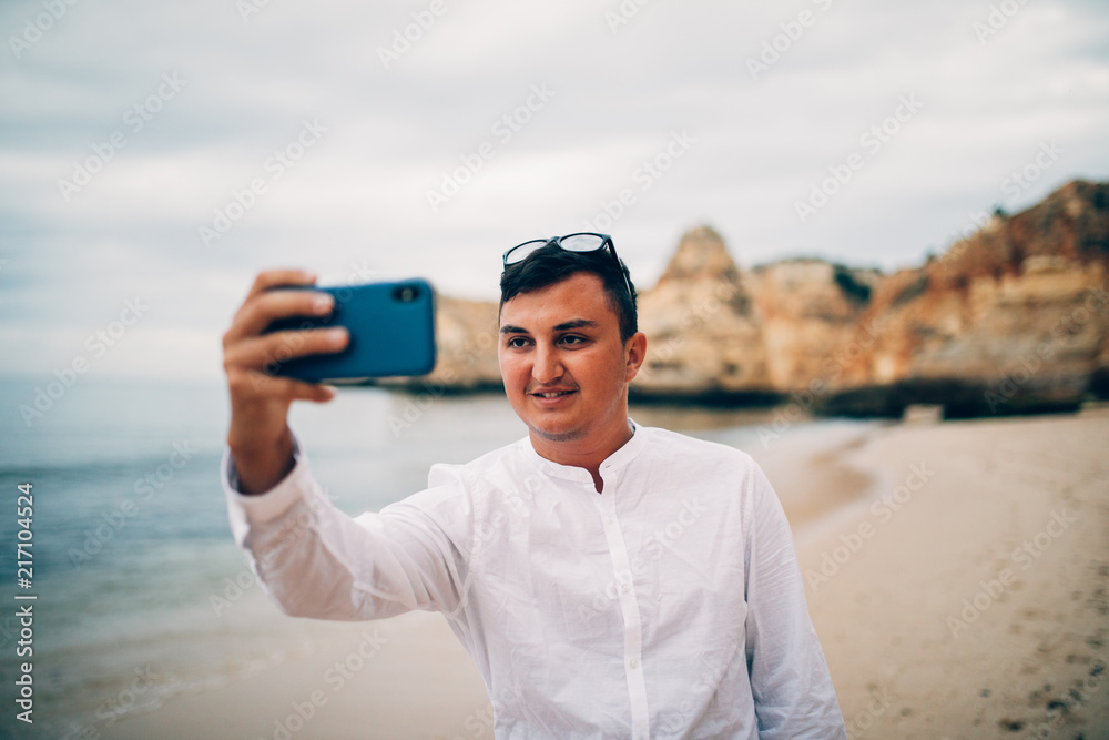 Happy young man in white shirt taking selfie with mobile phone while on vacation at the sea