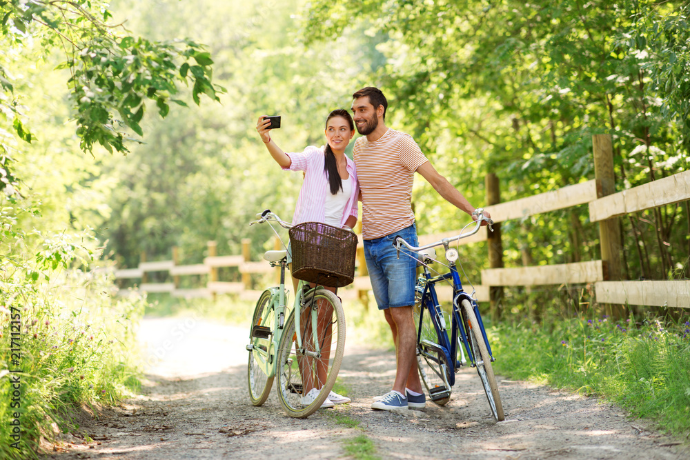 people, technology and lifestyle concept - happy couple with bicycles taking selfie by smartphone at summer park