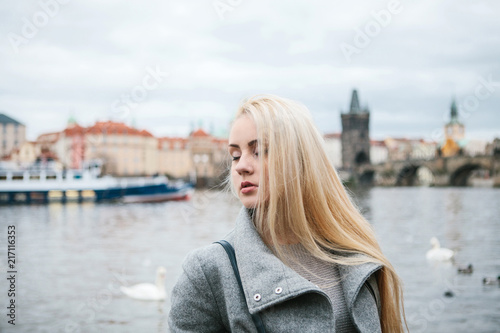 Fashionable blonde with long hair in a coat near Charles Bridge in Prague. Beautiful young woman outdoors.