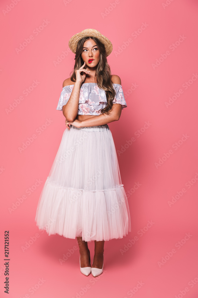 Full length portrait of brunette surprised woman 20s wearing straw hat and fluffy skirt thinking and touching chin with brooding glance, isolated over pink background Full length portrait of brunette surprised woman 20s wearing straw hat and fluffy skirt thinking and touching chin with brooding glance, isolated over pink background