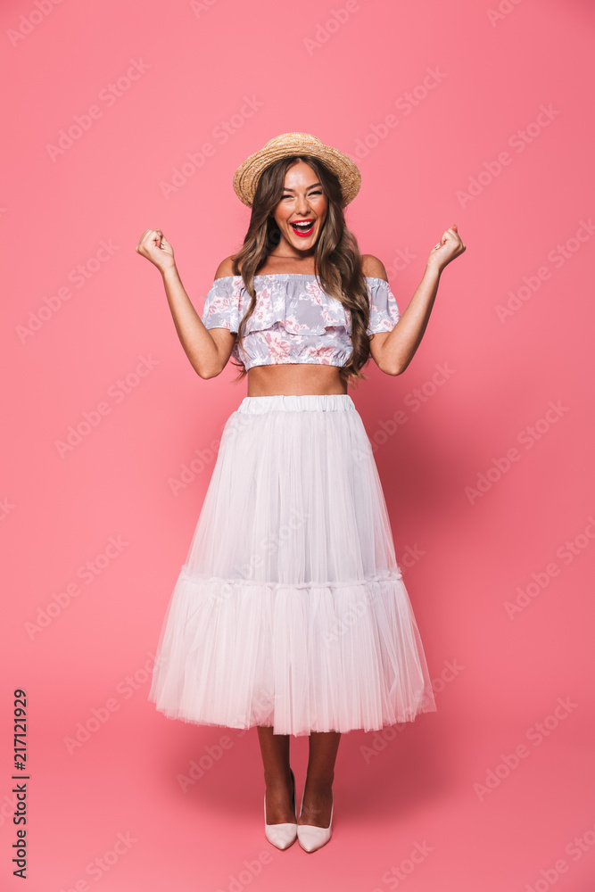 Naklejka premium Full length portrait of beautiful excited woman 20s wearing straw hat and fluffy skirt laughing while clenching fists like winner, isolated over pink background in studio