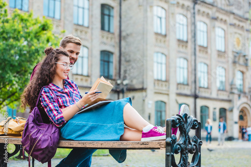 smiling young woman with book and notebook leaning on boyfriend on wooden bench in park
