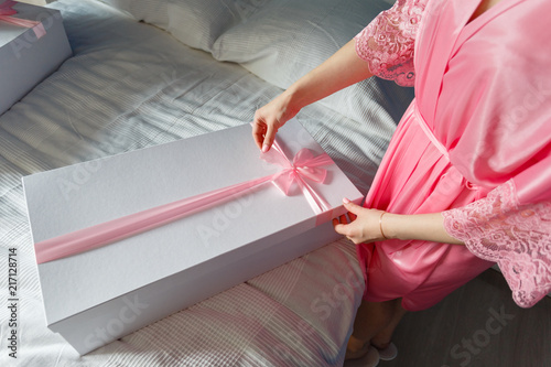 A young girl in a pink robe opens a white box with a bow with a new pink dress inside, unleashes a bow on the gift box. Surprise or gift on the bed in the bedroom.