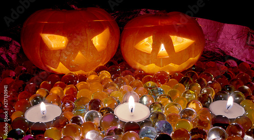 Two orange glowing carved pumpkin heads for the holiday of Helloon, surrounded by colorful jellied balls, with three candles, lie on a table on a dark background.