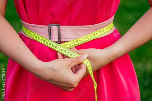 woman dressmaker seamstress measures the body with a measuring tape in a pink dress with belt in summer park. diet separate food on outdoors
