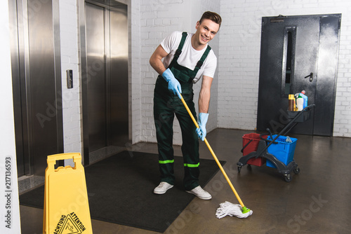 Handsome young janitor mopping floor and smiling at camera photo