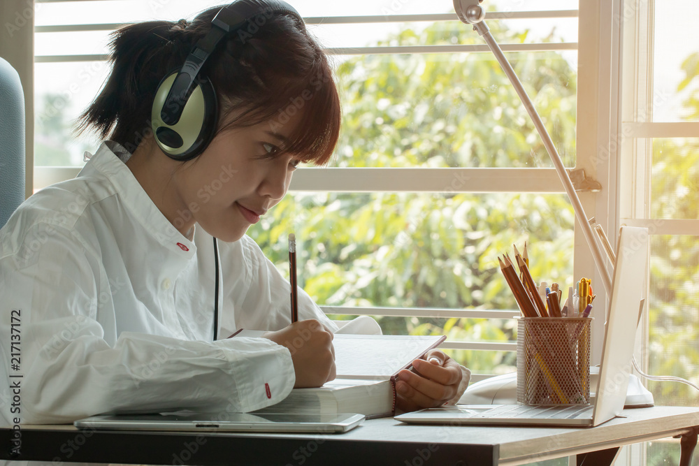 Student learning online study concept: Beautiful Asian girl listening with Headphones and laptop, sitting smile writing notes in textbook at her desk in home for e-learning in educational technology Student learning online study concept: Beautiful Asian girl listening with Headphones and laptop, sitting smile writing notes in textbook at her desk in home for e-learning in educational technology