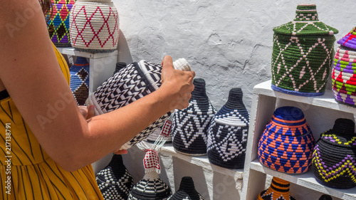 Tourist woman shopping moroccan handcraft souvenirs on the streets of the Medina (Old Town) of a Morocco village. Bright-colored weaved bottles and baskets.