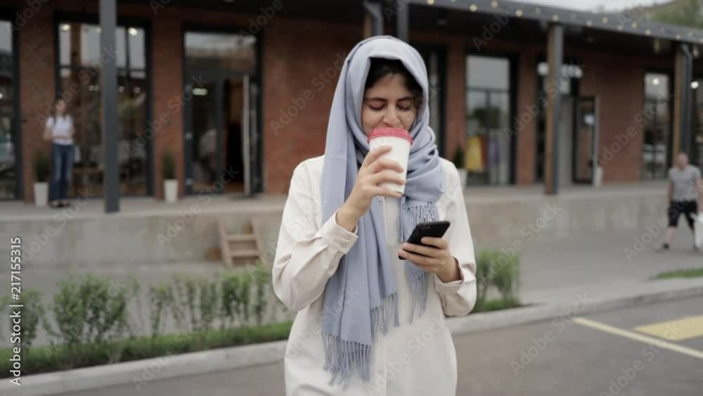 Attractive young woman in hijab walking, drinking coffee and looking at her smartphone screen. Tracking real time medium shot Attractive young woman in hijab walking, drinking coffee and looking at her smartphone screen. Tracking real time medium shot