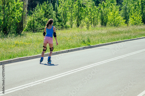 attractive young athletic slim brunette woman in pink shorts and blue top with protection elbow pads and knee pads rides on roller skates in the park