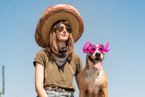 Beautiful girl in mexican hat dressed up as bandit of gangster with dog in cool sunglasses. Female person in sombrero hat and bandana posing with puppy as mexico festive symbol or for halloween © Photoboyko