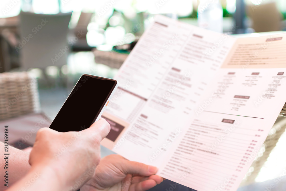 Person's hands holding menu and mobile phone in a restaurant. A man in a cafe ordering food while sitting at table. Mockup for smartphone screen Person's hands holding menu and mobile phone in a restaurant. A man in a cafe ordering food while sitting at table. Mockup for smartphone screen