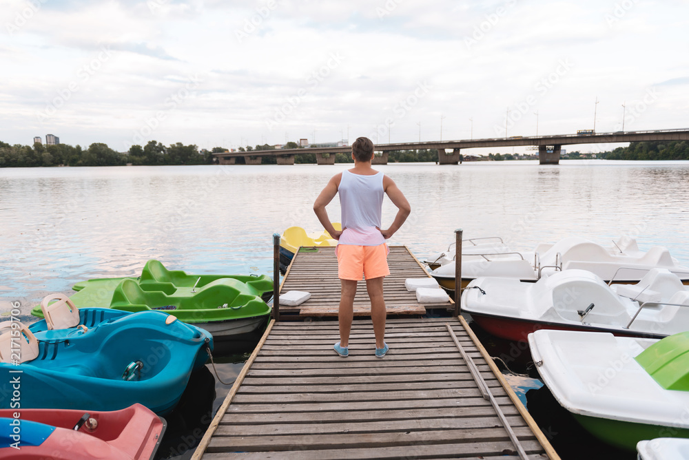 Near river. Good-looking muscle man wearing white shirt and orange shorts standing near river