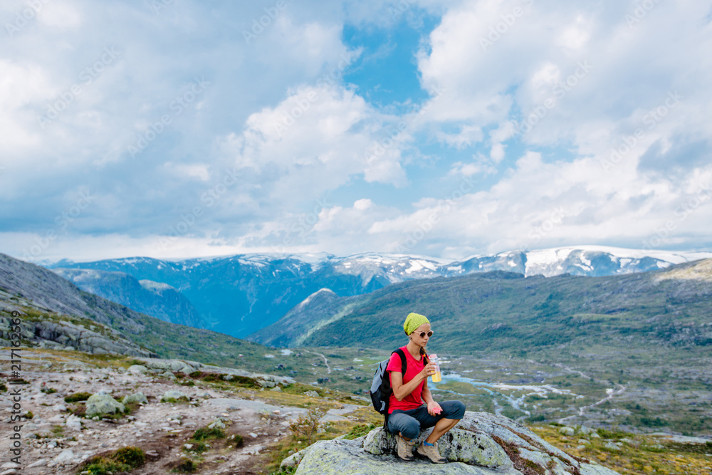 Travel in Norway, woman hiker in bright sportswear with backpack sitting on big stone and drinking water over scandinavian summer view.