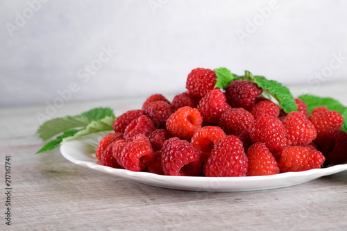  Red fresh raspberries on white rustic wood background. Plate with natural ripe organic berries with green leaves.flat lay with copy space