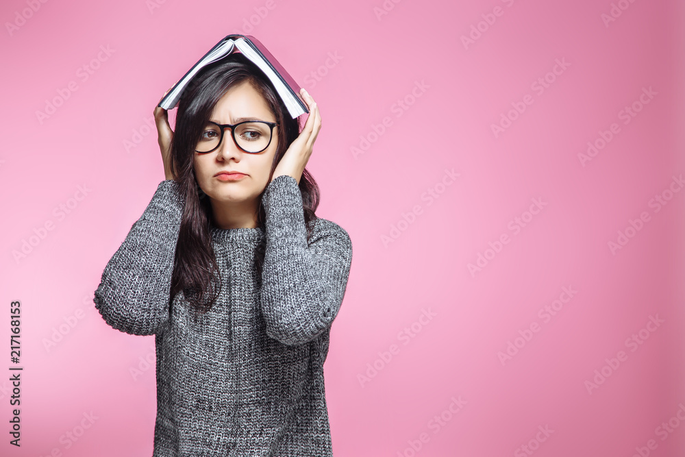 Sad teen student with book on head on pink background.