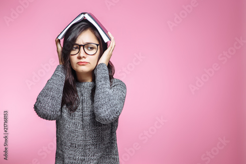 Sad teen student with book on head on pink background.