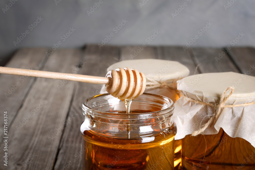 Pouring aromatic honey into jar, closeup. Honey in glass jars and honeycombs wax on wooden background. Wooden stick , instruments