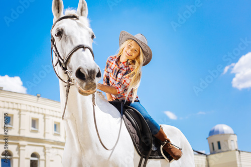 Touching horse. Beaming stylish schoolgirl touching her white racing horse while sitting on him